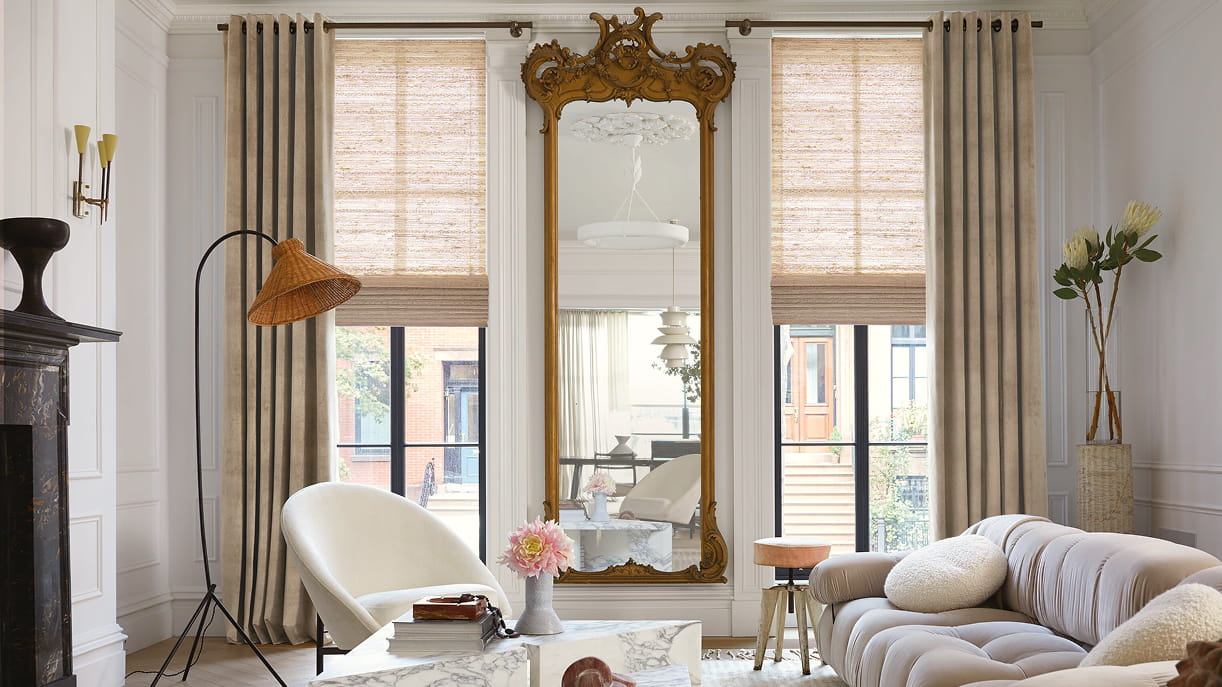 A luxe living room with an ornate mirror and Velvet grommet drapery in Antique layered with Cove woven wood shades in Ash.