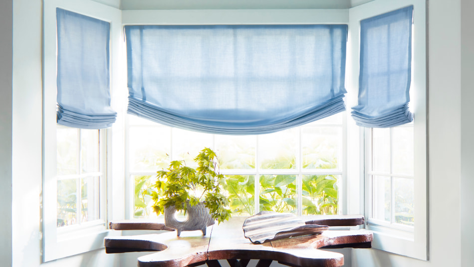 A box bay window with a wooden table in front features relaxed roman shades in blue Holland &amp; Sherry Andes fabric.