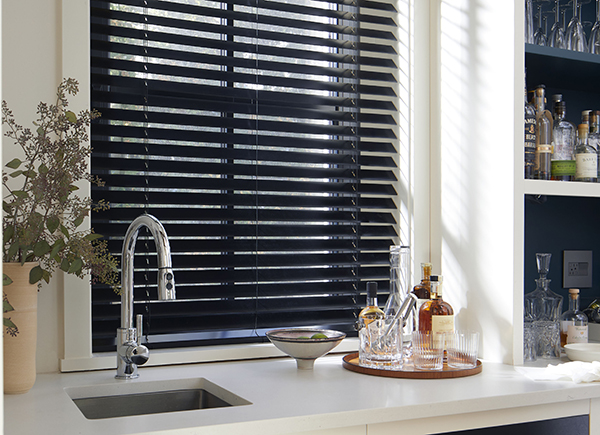 A window featuring 2 inch wood blinds over a small sink in a bar area with dark shelves holding various bottles and glasses