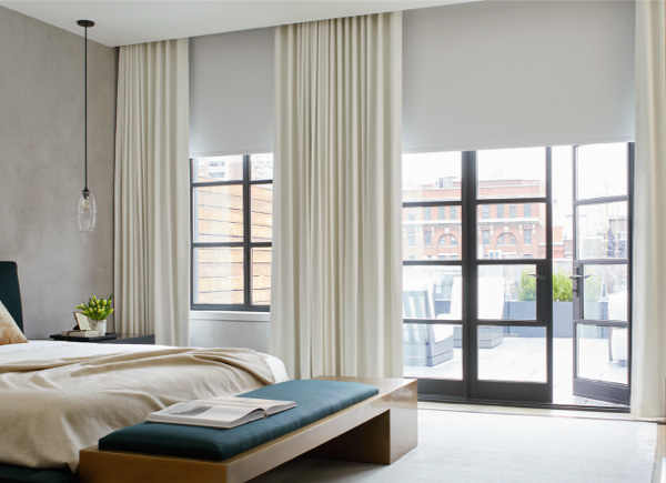 A patio door with large windows featuring blackout roller shades layered under drapery in a bedroom with a wooden bench