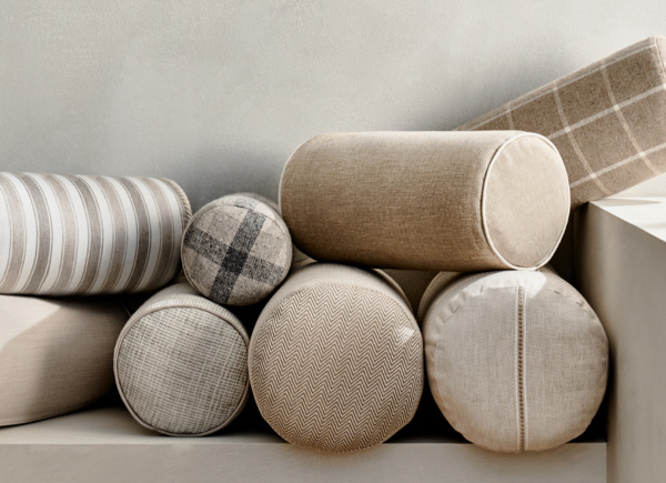 Multiple bolster pillows in neutral colored fabrics with various trim options stacked against a white wall