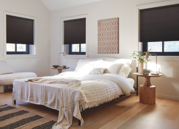 Three windows with black frames featuring cellular blackout shades in a bedroom with white walls, bedding and wooden tables