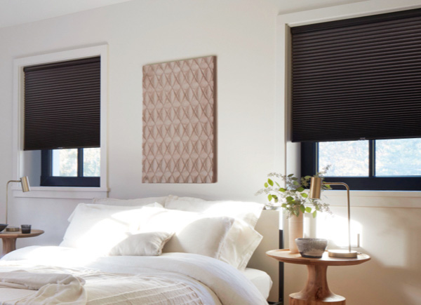 Two windows featuring cellular shades in single cell bo midnight in a bedroom with an all white bed and wooden night stands
