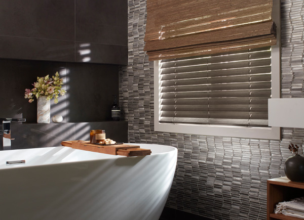A window featuring faux wood blinds layered under waterfall woven wood shades in a Bathroom with tiled walls and a bathtub