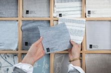 A pair of hands holding swatch squares over a opened showroom swatch drawer featuring various window treatment swatches