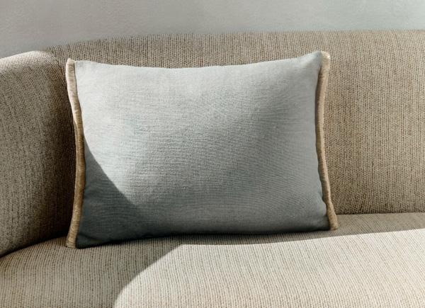 A singular rectangular pillow with fringe placed on a neutral couch against a white wall 