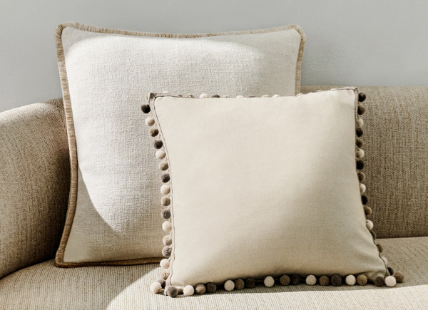 Two square pillows with fringe positioned on a neutral couch