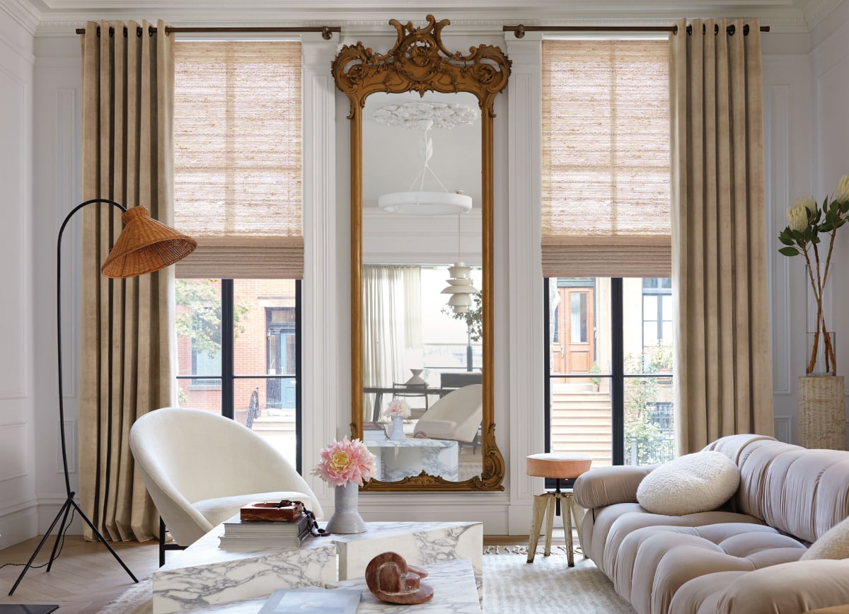 A luxe living room with an ornate mirror and Velvet grommet drapery in Antique layered with Cove woven wood shades in Ash.