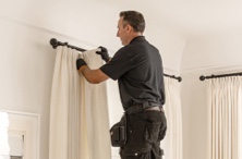 A Measure and Install professional on a ladder installing drapery wearing all black with black gloves