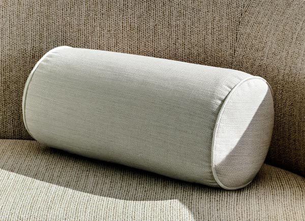 A sigular bolster pillow with piping in a linen blend material and ash color on a neutral colored couch