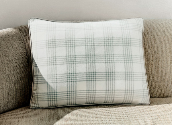 A singular rectangular pillow with piping placed on a neutral couch against a white wall 