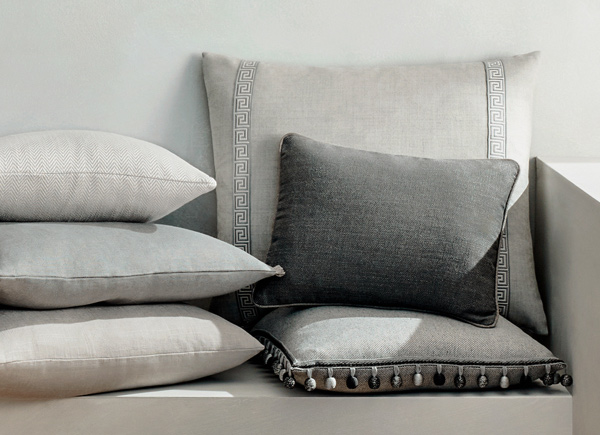 Multiple rectangular pillows in grey colored fabrics with various trim options stacked against a white wall