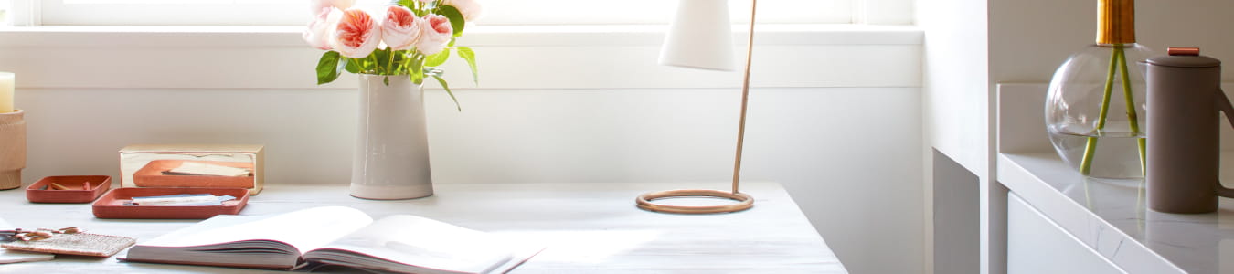 A home office with desk facing the window adorned with a floral arrangement, book and desk lamp.
