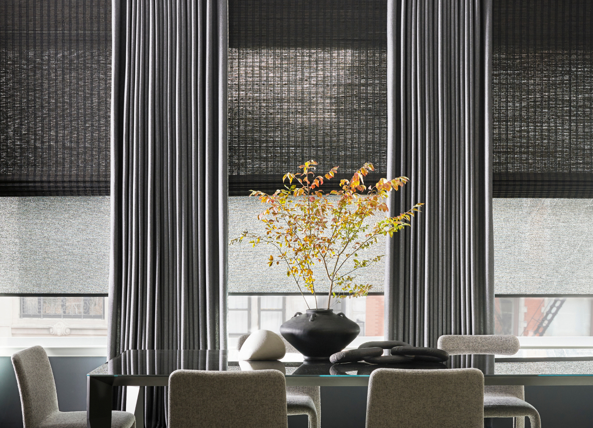 A dining room with a vase with plants has window treatments including: woven wood shades, roller shades and dark ripple fold