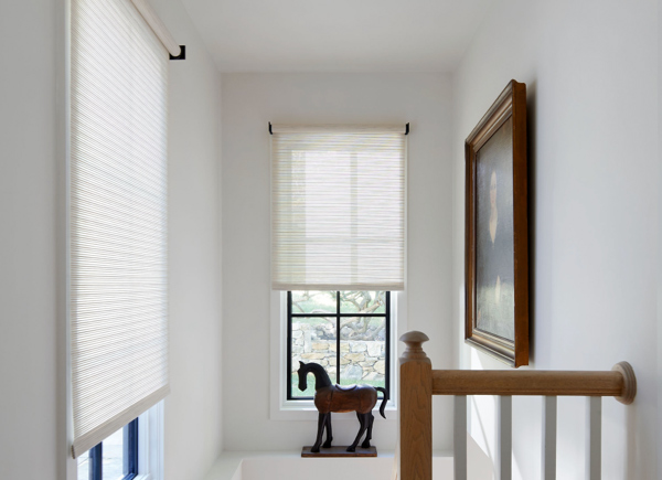 Two windows with black frames featuring roller shades in mesa verde sand over a staircase with a wood horse and painting