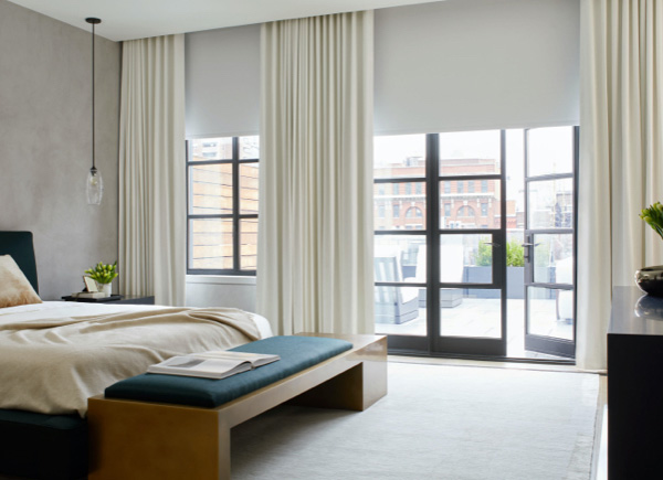 A patio door and large window featuring blackout roller shades layered under drapery in a bedroom with a wooden bench A patio door and large window featuring blackout roller shades layered under drapery in a bedroom with a wooden bench