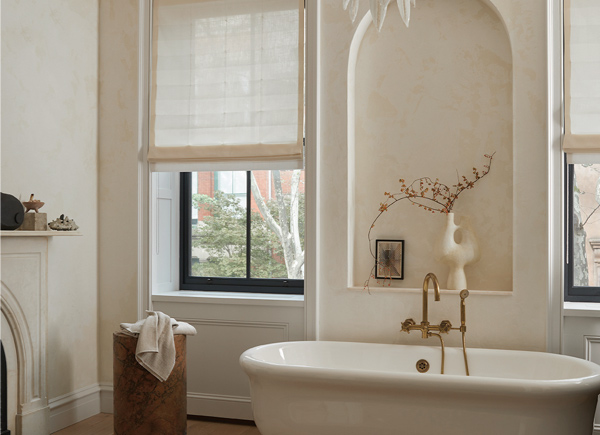 Two windows featuring roman shades in an all white Bathroom that has a freestanding bathtub with a gold faucet