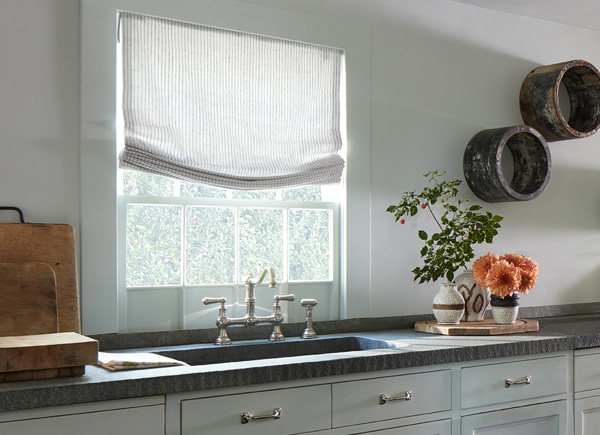 A window featuring relaxed roman shades in windsor stripe sage in a kitchen with a dark grey counter over white cabinets 