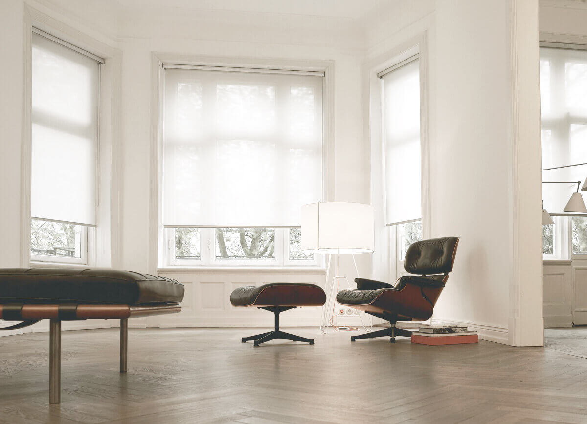 A bright open living room with a black chair and matching ottoman in front of a bay window dressed with white solar shades.