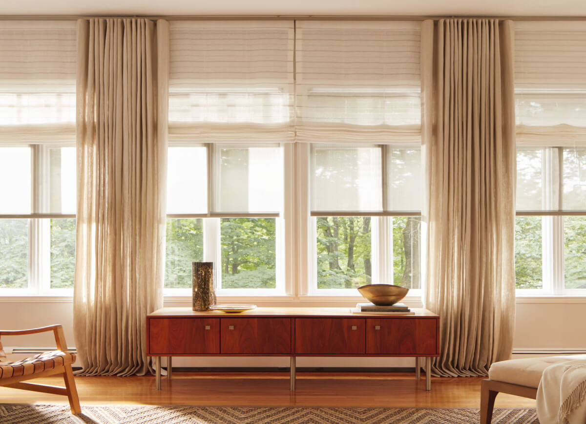 Living room with warm hued wood decor and a wall of windows layered with Solar Shades, Roman Shades and Ripple Fold Drapery.