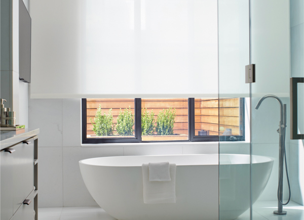 A large window featuring light filtering solar shades in white in an all white bathroom with a white freestanding bathtub