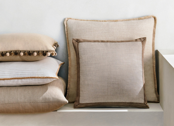 Multiple square pillows in neutral colored fabrics with various trim options stacked against a white wall