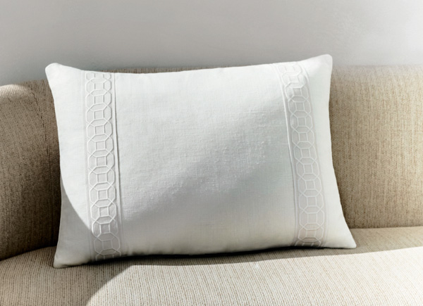 A singular rectangular pillow with tape border placed on a neutral couch against a white wall 
