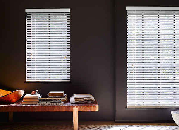 Two windows in various sizes featuring 2 inch wood blinds in a living room with dark walls and a leather lattice bench