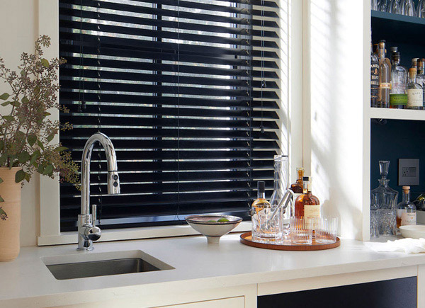 A window featuring 2 inch wood blinds over a small sink in a bar area with dark shelves holding various bottles and glasses