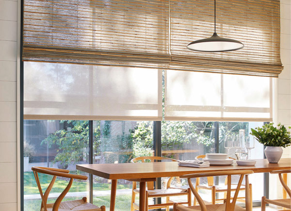 Floor to ceiling windows featuring woven wood shades layered over solar shades in a Dining Room with a wood table and chairs