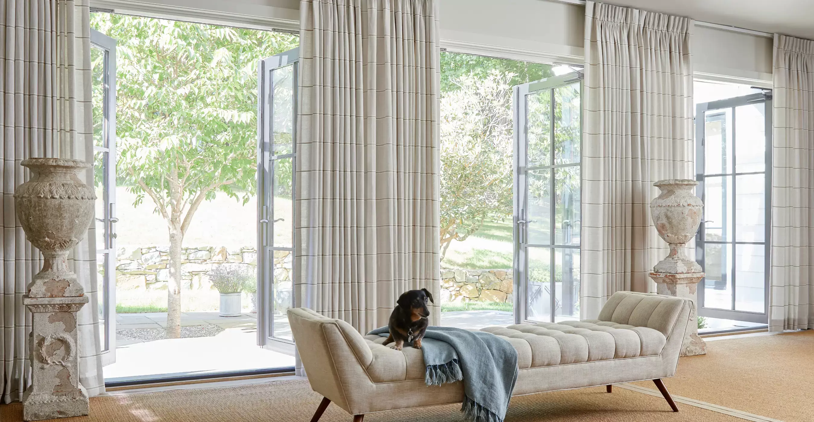 A traditional living room has open French doors with Pinch Pleat Drapery made of Victoria Hagan Sankaty Stripe in Sand.