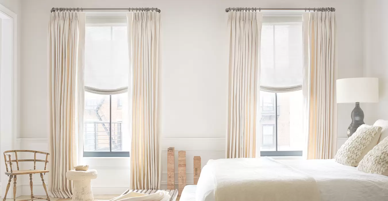 Two bedroom windows featuring Relaxed Roman Shades and Tailored Pleat Drapery in neutral tones with a bed made
