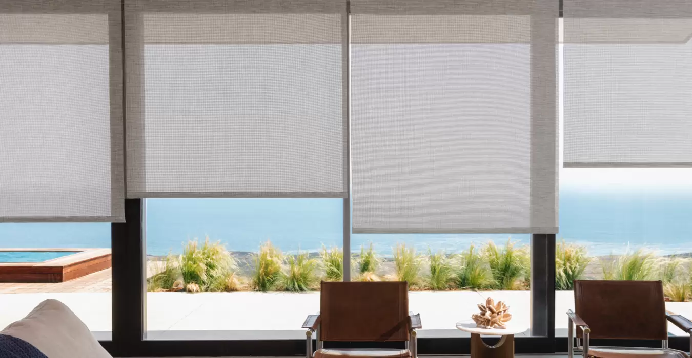 A beach house has huge windows with a view of the beach and Roller Shades made of Naomi in Honey pulled partially over them