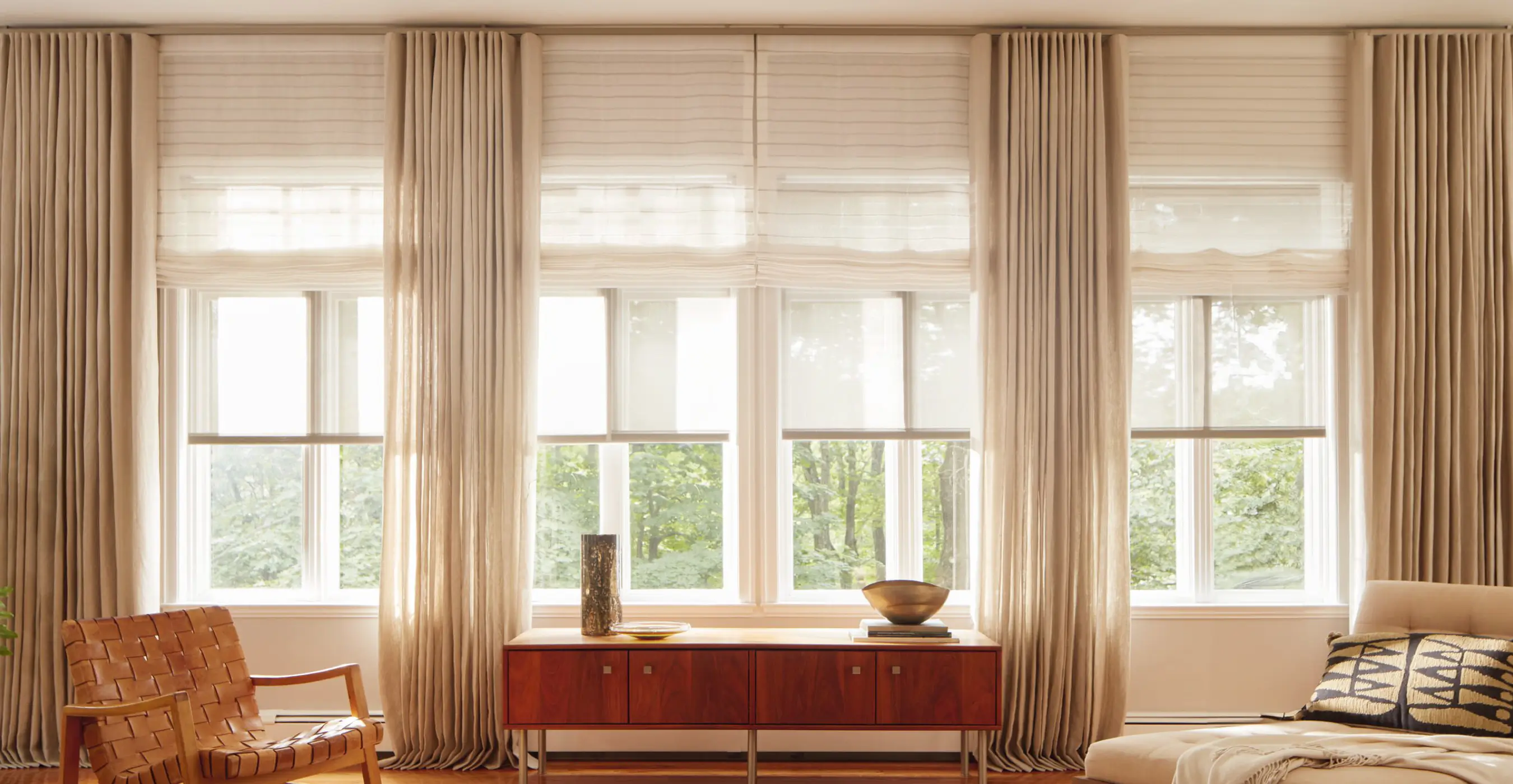 Living room with warm hued wood decor and a wall of windows layered with Solar Shades, Roman Shades and Ripple Fold Drapery.