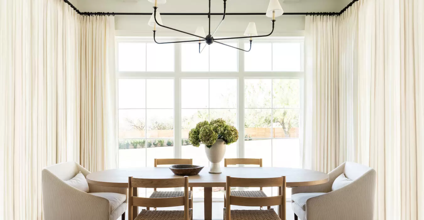 Lush Tailored Pleat Drapery made of Luxe Linen in Oyster hangs over large windows in a dining room with a light wood table