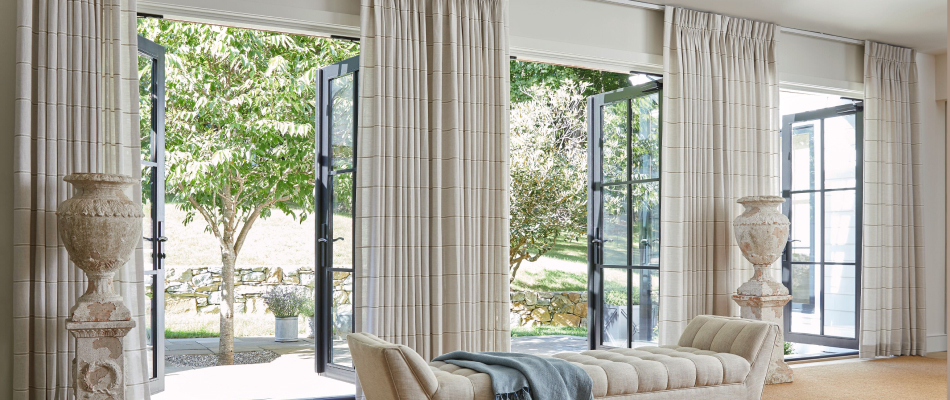 A traditional living room has open French doors with Pinch Pleat Drapery made of Victoria Hagan Sankaty Stripe in Sand