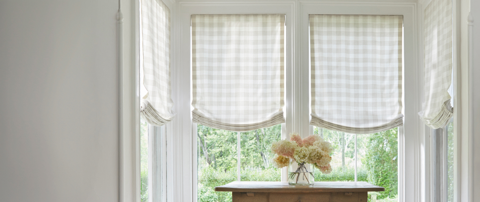 A quaint alcove has a table with flowers and Relaxed Roman Shades made of Holland & Sherry Emerson fabric over the windows