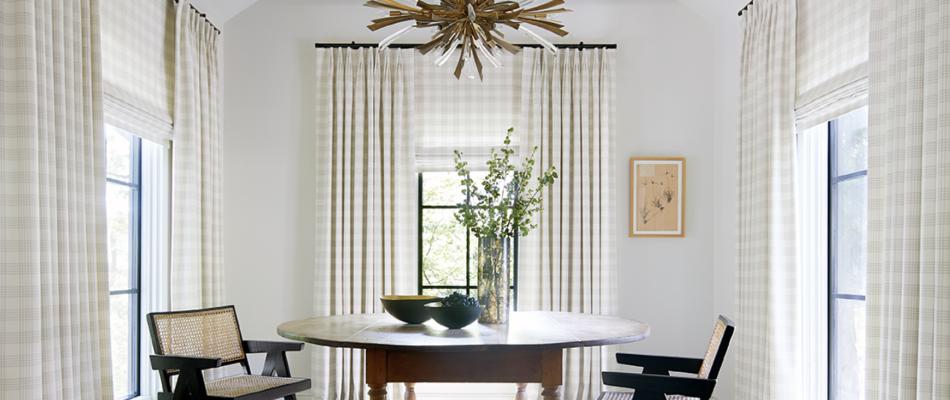 A dining room with a round wood table has Tailored Pleat Drapery and Flat Roman Shades made of Emerson in Shea