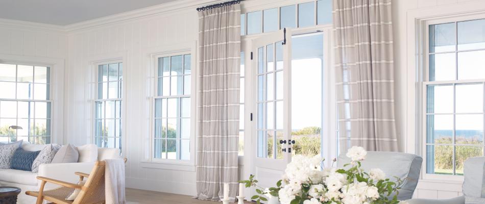 A breezy coastal room features Tailored Pleat Drapery made of Victoria Hagan Harbor Stripe in Sand over large glass doors