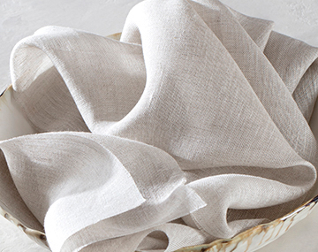 A swatch of Luxe Sheer Linen placed in a cream bowl on a white table top features a soft texture and off-white color