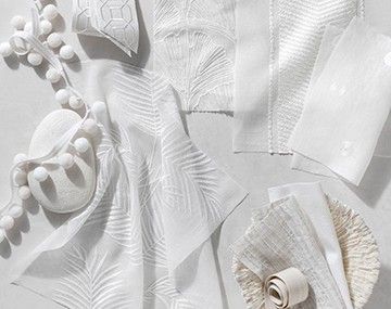 Delicate white fabrics with flowering embroidery sit on a white tabletop with tassels and a rock and seashell for texture