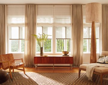 Tall windows featuring layered roller shades over roman shades and drapery in a living room with chairs and a side table