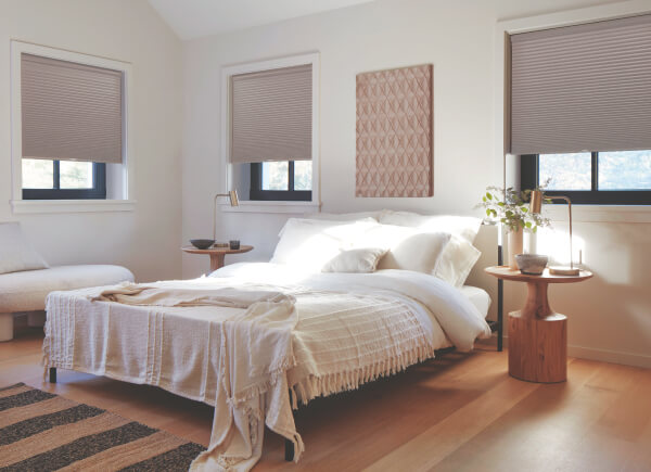 Blackout Cellular Shades in Ash hung over black framed windows in a bedroom with white bedding and brown side tables