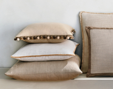 Square Pillows featuring neutral colored fabric and various piping styles are stacked invitingly against a white wall