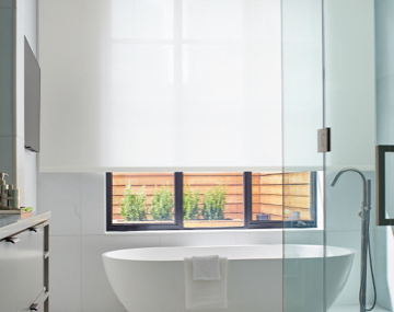 White Solar Shades soften light in a modern all-white bathroom with a large free stand tub and silver metal accents