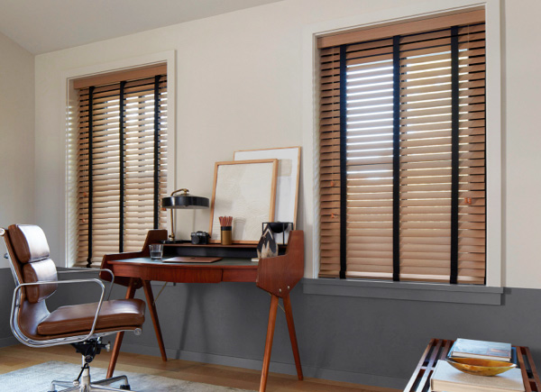 Two windows featuring wood blinds in an office with a brown chair and centered wood desk against grey and white walls