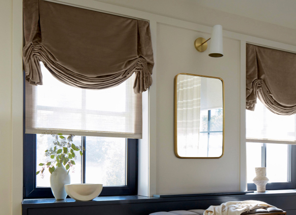 Two large windows with black frames featuring Tulip Roman Shades in velvet camel in a room with white vases and a mirror