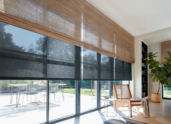 A large wall of windows featuring woven wood shades over roller shades in a room with a woven wood chair and large plant